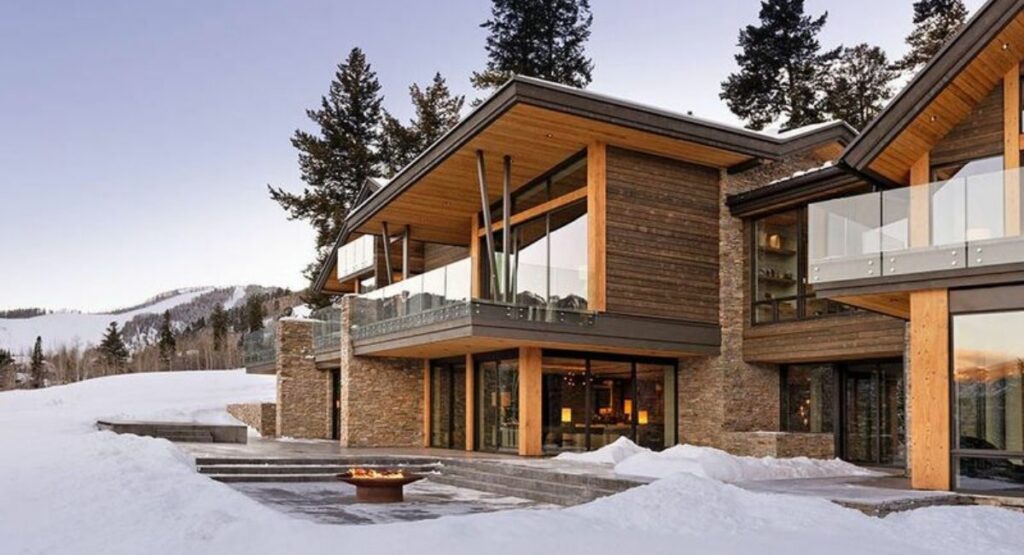 Modern  Aspen vacation rental with easy ski-in ski-out access, surrounded by snow. A fire pit on the patio creates a warm, inviting atmosphere amidst the winter setting.