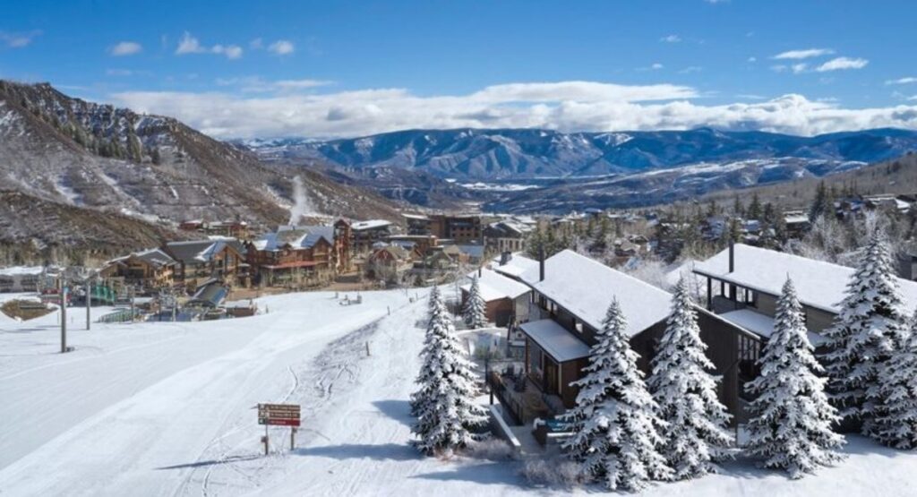 Snow-covered Aspen neighborhood with frosted pine trees. Majestic mountains and vibrant buildings under a clear blue sky enhance the serene winter scene.