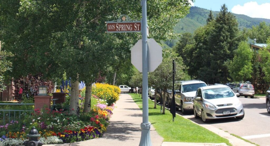 A sunny street scene with a "Spring St" sign, lush trees, and colorful flower beds lining the sidewalk. Parked cars and mountains are visible in the background in Aspen Core