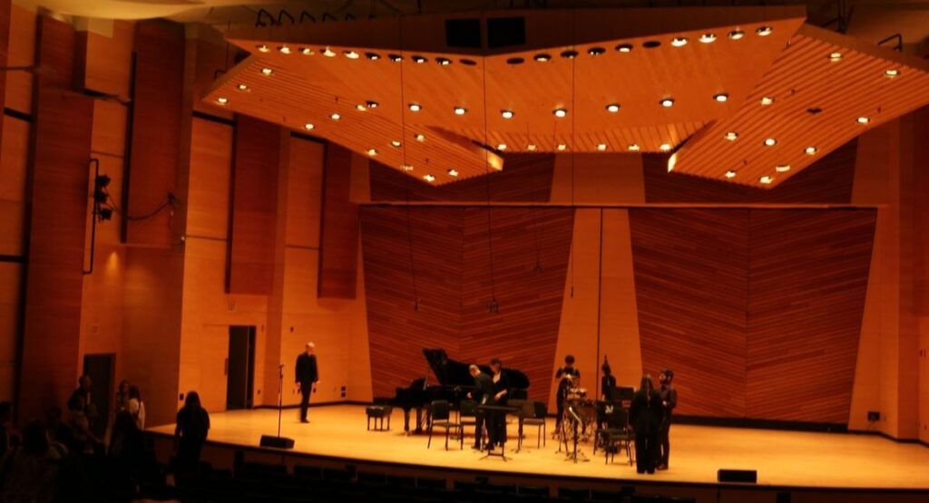 A warmly lit concert hall stage with a grand piano, chairs, music stands, and overhead lighting in Harris Concert Hall, Aspen